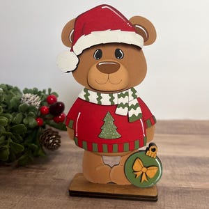 Puede incluir: Figura de oso navide&ntilde;o de madera que lleva un su&eacute;ter rojo con un &aacute;rbol de Navidad verde, un gorro de Pap&aacute; Noel rojo y una bufanda verde y blanca. El oso sostiene un adorno verde con un lazo dorado.