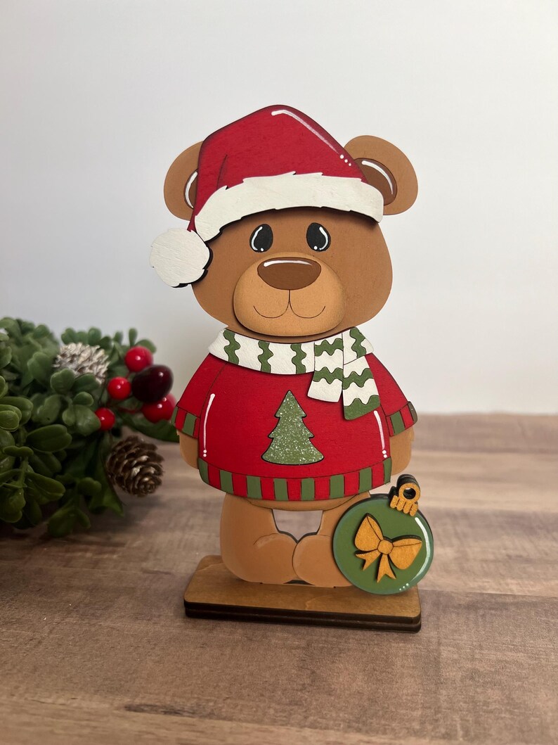 Puede incluir: Figura de oso navide&ntilde;o de madera que lleva un su&eacute;ter rojo con un &aacute;rbol de Navidad verde, un gorro de Pap&aacute; Noel rojo y una bufanda verde y blanca. El oso sostiene un adorno verde con un lazo dorado.