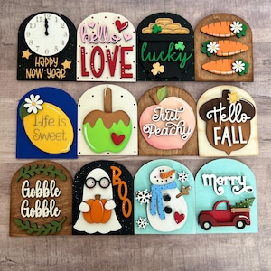 May include: A collection of decorative wooden signs with various themes. Designs include a clock, the words "hello LOVE", a pot of gold, carrots, a lemon, an apple, a peach, an acorn, the words "Gobble Gobble", a ghost, a snowman, and a red truck.
