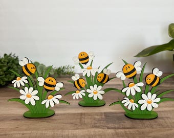 May include: Three wooden decorative pieces featuring bees, daisies, and green grass. Each piece has a green base, white daisies, and cartoon bees with black and yellow stripes. The background is a light wall and a wooden surface.