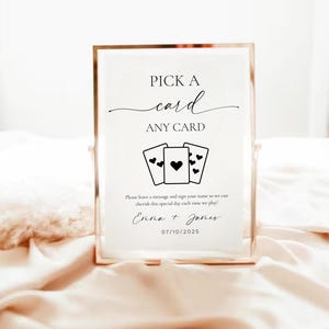 以下が含まれることがあります： 「PICK A card ANY CARD」という文字と、ハートのマークが描かれたトランプ4枚のグラフィックが書かれた、金色の枠のある白い看板。看板には、「Please leave a message and sign your name so we can cherish this special day each time we play! Emma + James 07/10/2025」という文字も書かれている。