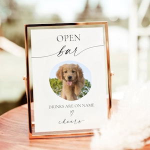 May include: White sign with black text that reads "Open bar" with a cursive font. A photo of a golden retriever puppy is in the center of the sign. The text below the photo reads "Drinks are on name" with a heart symbol and "cheers" in cursive font.