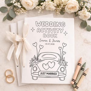 May include: A white wedding activity book with a black line drawing of a car featuring heart-shaped balloons, floral accents, and a licence plate reading JUST MARRIED. The cover includes the text WEDDING ACTIVITY BOOK, Emma & James, and 01/07/2028.