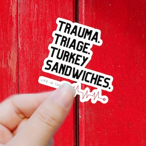 May include: White sticker with black text that reads "Trauma. Triage. Turkey Sandwiches." and a red heartbeat graphic below the text. The sticker is on a red surface.