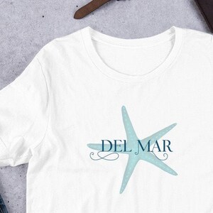 Peut inclure: T-shirt blanc avec une étoile de mer bleue et le texte "DEL MAR" imprimé sur le devant.
