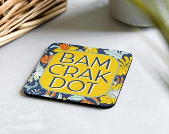 Könnte beinhalten: Quadratischer Untersetzer mit floralem Muster in Marineblau, Gelb, Orange und Weiß. Der Untersetzer trägt die Worte "BAM CRAK DOT" in einem gelben Rechteck. Der Untersetzer liegt auf einer hellgrauen Oberfläche.