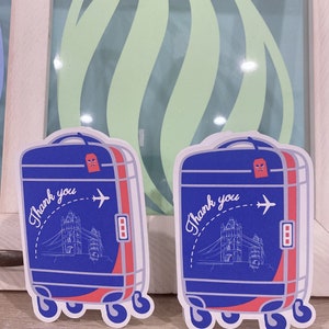 May include: Two blue and red suitcase-shaped stickers. Each sticker features a drawing of the Tower Bridge, an airplane, and the words "Thank you". The stickers have a white border and are on a wooden surface.