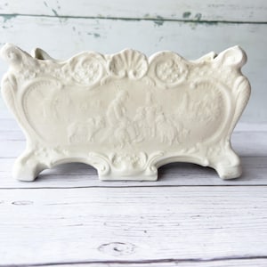 May include: Cream-coloured ceramic planter with a rectangular shape and ornate relief detailing. The planter features a central panel with a classical scene, supported by four feet and decorative scrollwork. The top edge has a scalloped design.