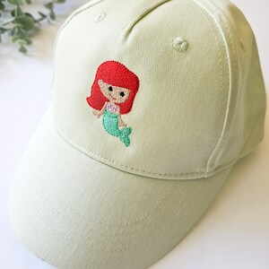 May include: A light green baseball cap with an embroidered mermaid design. The mermaid has red hair, a pink seashell, and a green tail.