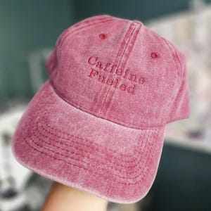 May include: A washed-out red baseball cap with a six-panel construction and a curved brim. The front of the cap features the embroidered text "Caffeine Fueled" in a matching red thread. The fabric has a distressed, vintage-style texture.