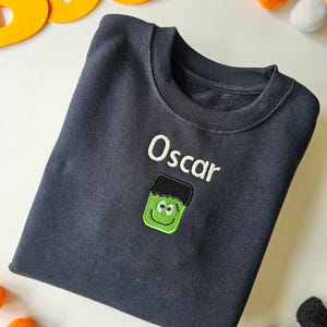 Black crew neck sweatshirt, embroidered with a green monster and personalised with childs name in glow in the dark thread. Machine embroidered in the UK
