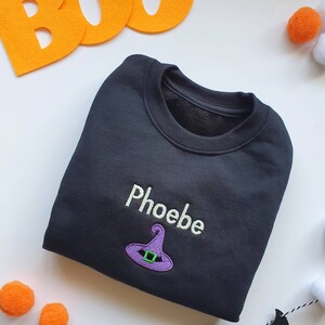 Black crew neck sweatshirt, embroidered with a purple witches hat and personalised with childs name in glow in the dark thread. Machine embroidered in the UK