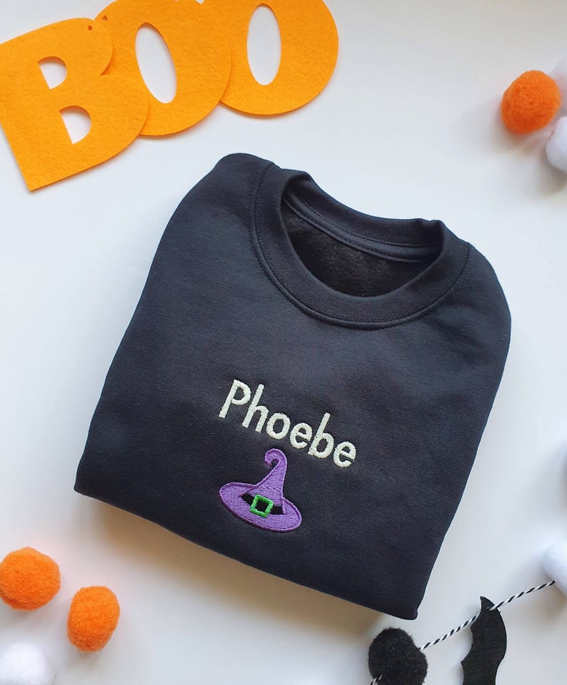 Black crew neck sweatshirt, embroidered with a purple witches hat and personalised with childs name in glow in the dark thread. Machine embroidered in the UK
