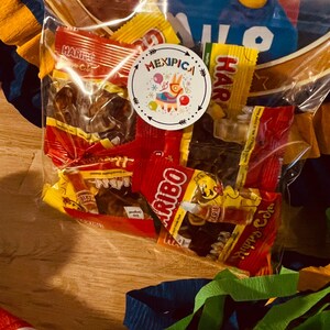 May include: A clear plastic bag filled with various colorful candy wrappers. The bag is sitting on a wooden surface. The candy wrappers are for Haribo,  Mexican candy, and other brands. The bag is labeled "Mexipica".