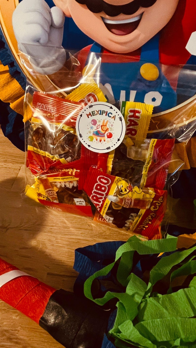 May include: A clear plastic bag filled with various colorful candy wrappers. The bag is sitting on a wooden surface. The candy wrappers are for Haribo,  Mexican candy, and other brands. The bag is labeled "Mexipica".