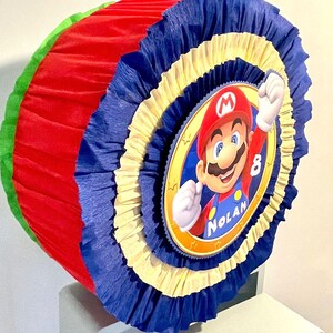 May include: A red, yellow, blue, and green pinata with a circular image of Mario from the Super Mario video game series. The pinata has the name "Nolan" and the number "8" on it.