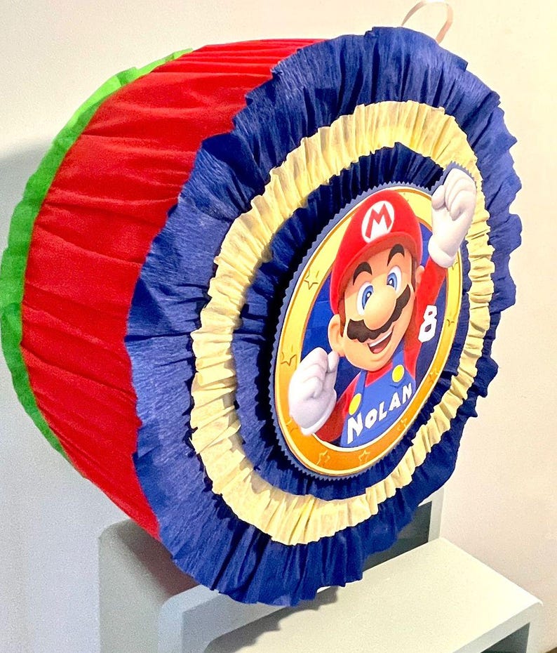 May include: A red, yellow, blue, and green pinata with a circular image of Mario from the Super Mario video game series. The pinata has the name "Nolan" and the number "8" on it.