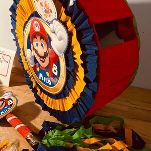 May include: A red and blue pinata with a Mario character on the front. The pinata is decorated with yellow and blue tissue paper. The pinata has a string attached to the top and says "Milo 8" on the front.