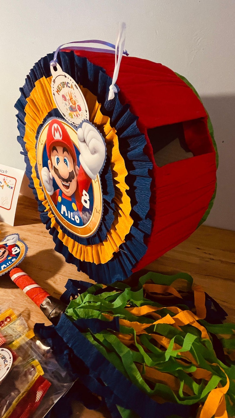 May include: A red and blue pinata with a Mario character on the front. The pinata is decorated with yellow and blue tissue paper. The pinata has a string attached to the top and says "Milo 8" on the front.