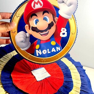 May include: A red, blue, and yellow paper pinata with a round, gold-rimmed image of Mario from the Super Mario Bros. video game series. The image includes the text "Nolan" and the number "8".