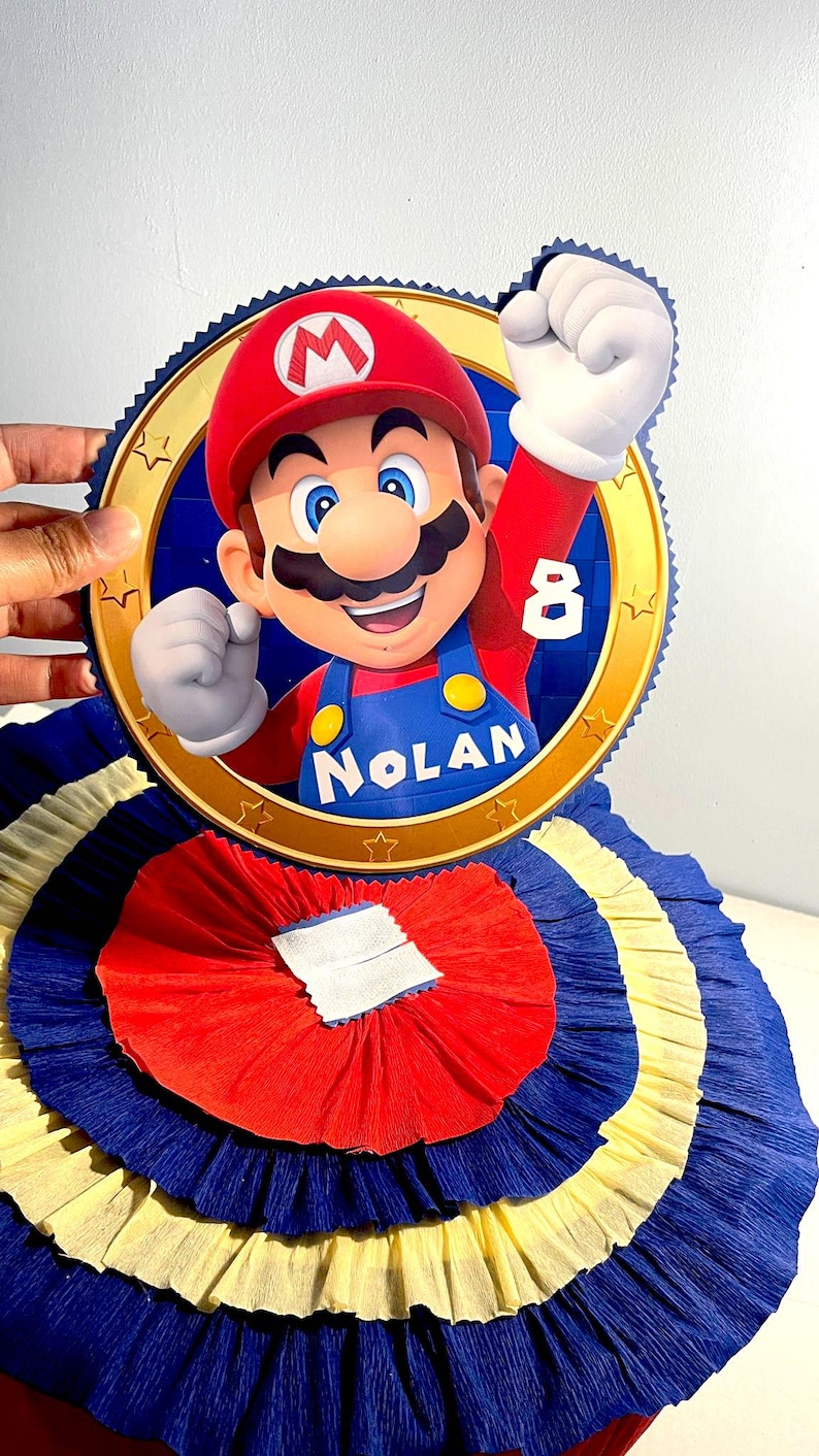 May include: A red, blue, and yellow paper pinata with a round, gold-rimmed image of Mario from the Super Mario Bros. video game series. The image includes the text "Nolan" and the number "8".