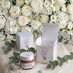Peut inclure: Des cadeaux de baptême pour bébé, comprenant de petites boîtes cadeaux et un pot de pâte à tartiner, avec le nom "Louca" imprimé. Les boîtes et le pot sont décorés d'un ours en peluche et de ballons. Fond de fleurs blanches.