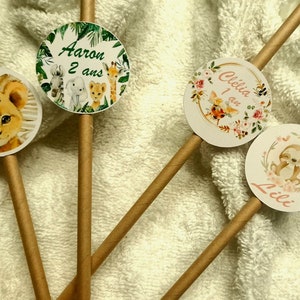 Peut inclure: Quatre pailles en papier brun avec des autocollants ronds en papier blanc attachés. Les autocollants présentent des illustrations d'animaux et du texte qui se lit "Ediyh 1 an", "Aaron 2 ans", "Celia 1 an" et "Lili".