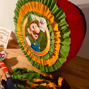 May include: A red, green, and yellow pinata shaped like a circle with a picture of a cartoon character wearing a green hat and green overalls. The character is holding a fist in the air. The pinata is decorated with green and yellow tissue paper.