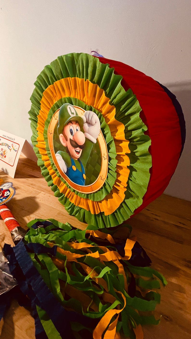 May include: A red, green, and yellow pinata shaped like a circle with a picture of a cartoon character wearing a green hat and green overalls. The character is holding a fist in the air. The pinata is decorated with green and yellow tissue paper.
