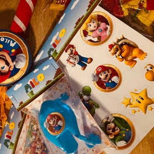 May include: A colorful Mario-themed party decoration set with a pinata, banners, and stickers. The pinata features a large image of Mario with the name "Milo" on it. The banners have the name "Milo" printed on them and feature a Mario-themed design. The stickers feature various characters from the Mario franchise, including Mario, Luigi, Bowser, and Toad.