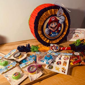 May include: A colorful pinata shaped like a character from the video game Super Mario Bros. The pinata is decorated with red, yellow, blue, and black colors. The character is wearing a red hat and overalls. The pinata is surrounded by party favors, including small toys, candy, and confetti. The text "Milo" is written on the pinata.