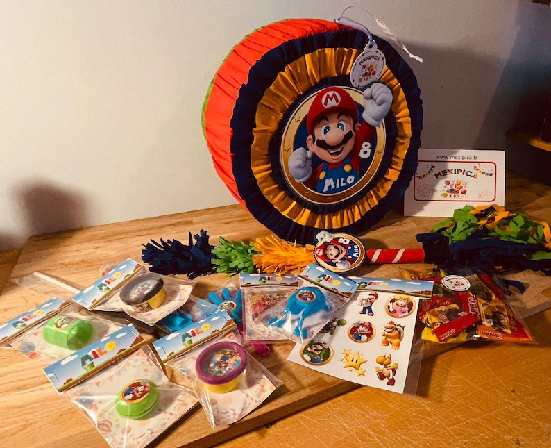 May include: A colorful pinata shaped like a character from the video game Super Mario Bros. The pinata is decorated with red, yellow, blue, and black colors. The character is wearing a red hat and overalls. The pinata is surrounded by party favors, including small toys, candy, and confetti. The text "Milo" is written on the pinata.