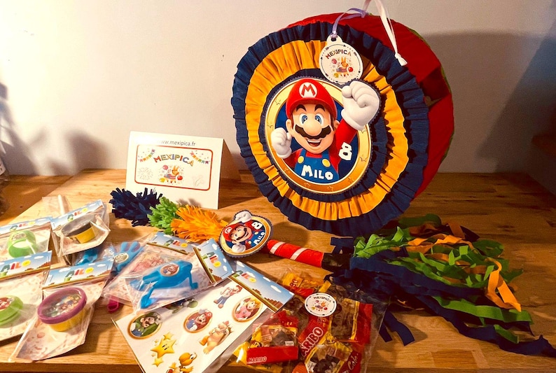 May include: A colorful Mario-themed pinata with a blue, yellow, and red design. The pinata features a picture of Mario with the name "Milo" written below. The pinata is surrounded by party favors, including small toys, candy, and a stick for hitting the pinata.