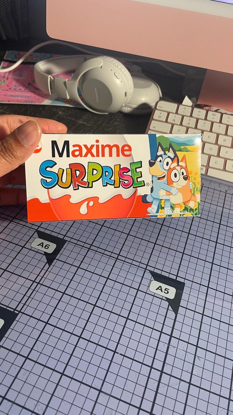 Pu&ograve; includere: Una scatola rettangolare di caramelle Maxime Surprise. La scatola presenta le parole "Maxime SURPRISE" in lettere colorate. Il lato destro della scatola presenta un'immagine dei cartoni animati di Bluey e Bingo. La scatola &egrave; tenuta da una mano.