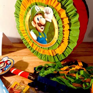 May include: A green, yellow, and red pinata shaped like a circle with a picture of Luigi from the Super Mario Bros. video game series. The pinata has a string attached to it and is surrounded by colorful streamers.