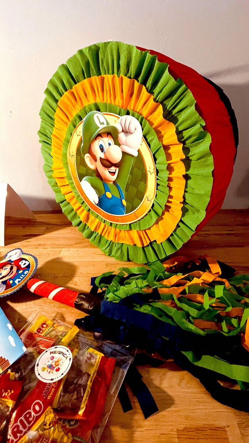 May include: A green, yellow, and red pinata shaped like a circle with a picture of Luigi from the Super Mario Bros. video game series. The pinata has a string attached to it and is surrounded by colorful streamers.