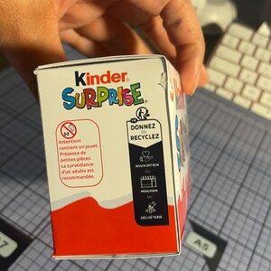 Pu&ograve; includere: Una scatola di uova Kinder Sorpresa, con il marchio in rosso e la parola &laquo;SURPRISE&raquo; in lettere colorate. La scatola presenta testo in francese che avverte della presenza di piccole parti e della necessit&agrave; di supervisione da parte di un adulto. Include anche simboli di riciclaggio.