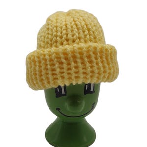 Könnte beinhalten: Ein grüner Eierbecher mit einem lächelnden Gesicht, bedeckt mit einer kleinen, gelben Strickmütze. Die Mütze hat einen umgeschlagenen Rand und ein strukturiertes Design. Der Eierbecher ist ein dekorativer Artikel.