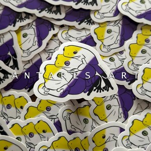 May include: A die-cut sticker featuring two stacked frogs colored in yellow, white, and purple stripes. The top frog rests on the back of the bottom frog, both depicted with black outlines. The text ANTARES ART is printed in white across the center.