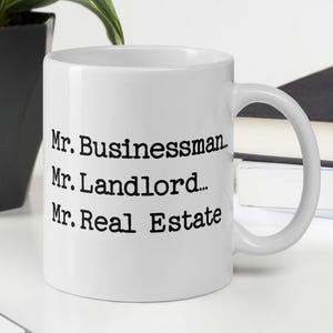 May include: White ceramic mug with the text "Mr. Businessman, Mr. Landlord... Mr. Real Estate" in black. The mug is on a white surface with a keyboard, a plant, and a book in the background.