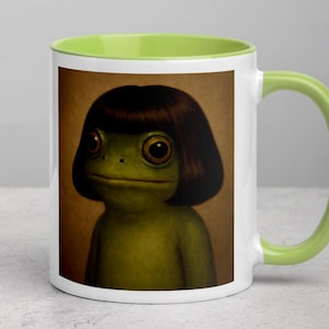 Puede incluir: Taza de cerámica blanca con interior y asa verdes. La taza presenta una ilustración caprichosa de una rana verde con cabello oscuro. La rana tiene ojos grandes y expresivos y una expresión neutra. El fondo es de un tono cálido y neutro.