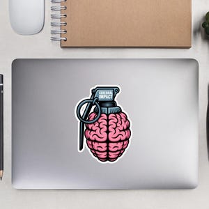 May include: A sticker featuring a pink brain shaped like a grenade with the text "Cerebral Impact" in a blue rectangle. The sticker is on a laptop, with a mouse, notebook, pen, glasses, and small plants nearby.