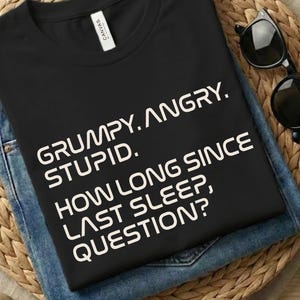 Op de afbeelding: Zwart T-shirt met de tekst "GRUMPY. ANGRY. STUPID. HOW LONG SINCE LAST SLEEP, QUESTION?" op een blauwe jeans. Het shirt en de jeans liggen op een geweven mat met zonnebril en stenen.