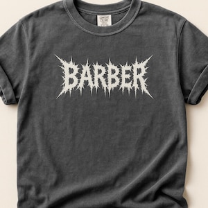 May include: Dark grey t-shirt with rolled sleeves. The word "BARBER" is printed in a spiky, white font across the chest. The shirt has a vintage, worn appearance.