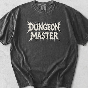 May include: A charcoal gray short-sleeve t-shirt with a vintage-washed finish. The center of the chest features the text DUNGEON MASTER printed in a white, jagged, metal-style font. The word PEPPER is printed in the top left corner of the image.