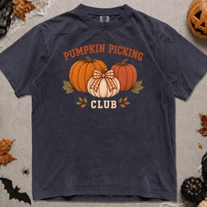May include: A black short-sleeve t-shirt featuring a graphic of three pumpkins with a plaid bow on the centre pumpkin. The text PUMPKIN PICKING CLUB is printed in orange above the pumpkins. The shirt has a crew neckline and a relaxed fit.