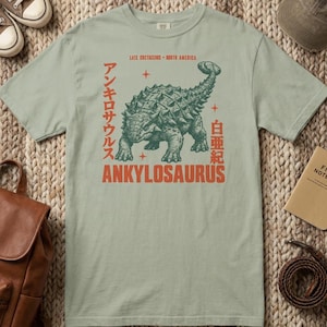 Puede incluir: Una camiseta de manga corta color verde salvia con un gráfico en verde oscuro de un dinosaurio Ankylosaurus. El estampado incluye el texto "LATE CRETACEOUS - NORTH AMERICA", "ANKYLOSAURUS" y caracteres japoneses, junto a pequeñas estrellas.