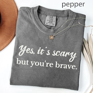 May include: A grey t-shirt with the text "Yes, it's scary but you're brave" in white script. A gold necklace with a small pendant is draped across the shirt. The shirt is folded, and a brown hat is partially visible.