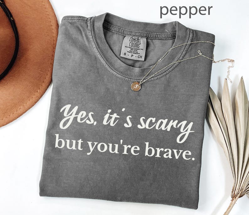 May include: A gray t-shirt with the text "Yes, it's scary but you're brave" in white script. A gold necklace with a small pendant is draped across the shirt. The shirt is folded, and a brown hat is partially visible.