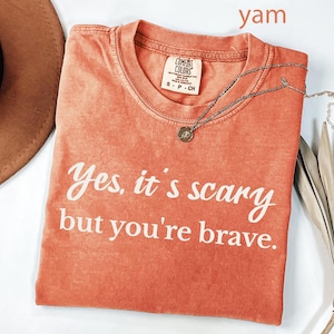May include: A coral-colored Comfort Colors t-shirt with the text "Yes, it's scary but you're brave" in white. A silver necklace with a small pendant rests on the shirt. A brown hat and dried leaves are in the background.